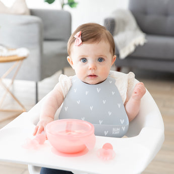 A baby in a gray bib with a bow sits in a high chair with a Bumkins Silicone First Feeding Set: Pink Jelly on the tray.