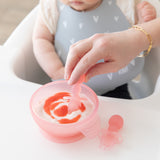 Close-up of an adult hand dipping a spoon into yogurt in a Bumkins Silicone First Feeding Set: Pink Jelly in front of a baby in a gray bib.