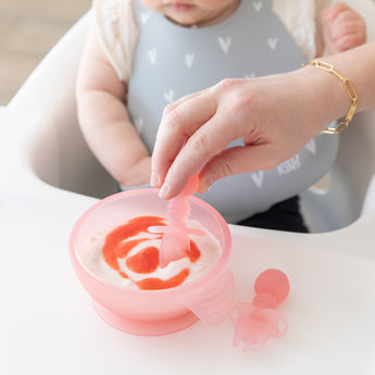 Close-up of an adult hand dipping a spoon into yogurt in a Bumkins Silicone First Feeding Set: Pink Jelly in front of a baby in a gray bib.