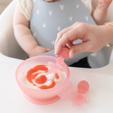Close-up of an adult hand using a Bumkins Silicone Chewtensils®: Pink Jelly to scoop yogurt from a pink bowl in front of a baby.
