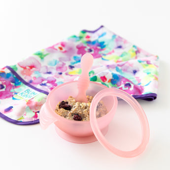 Bumkins Silicone First Feeding Set: Pink Jelly, filled with oatmeal and berries, sits next to a folded floral bib on a white background.