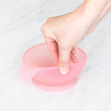 A hand holds a Bumkins Silicone First Feeding Set: Pink Jelly, demonstrating the suction cup bottom on a marble surface.