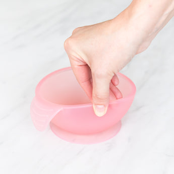 A hand holds a Bumkins Silicone First Feeding Set: Pink Jelly, demonstrating the suction cup bottom on a marble surface.