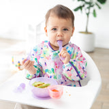 A baby with a colorful bib sits in a high chair using Bumkins Silicone First Feeding Set: Purple Jelly, surrounded by plants and toys.