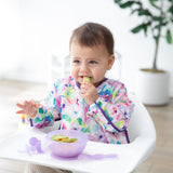 A baby in a floral bib sits in a high chair, nibbling from a Bumkins Silicone First Feeding Set: Purple Jelly by a potted plant.