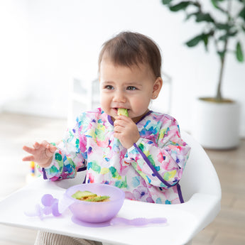 A baby in a floral bib sits in a high chair, nibbling from a Bumkins Silicone First Feeding Set: Purple Jelly by a potted plant.