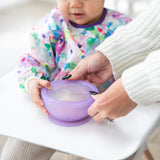 Adult hands guide a toddler with a Bumkins Silicone First Feeding Set: Purple Jelly at a high chair, wearing a colorful bib.