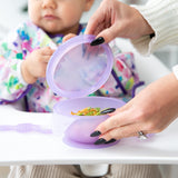 An adult opens a Bumkins Silicone First Feeding Set: Purple Jelly in front of a toddler in a floral bib, seated eagerly in a high chair.