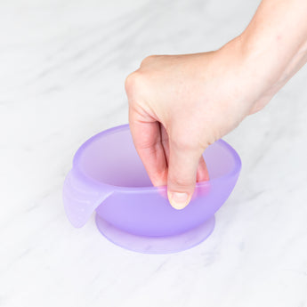 A hand holds a Bumkins Silicone First Feeding Set: Purple Jelly, demonstrating the suction cup bottom on a marble surface.