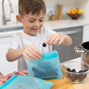 A smiling child in a kitchen places blackberries into a Bumkins Silicone Flat Reusable Bag 2 Pack, Blue, with a bowl nearby.