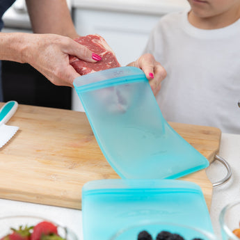 A person puts raw meat in a Bumkins Silicone Flat Reusable Bag 2 Pack, Blue as a child watches at a kitchen counter.
