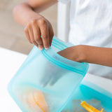 A hand puts orange slices into a Bumkins Silicone Flat Reusable Bag 2 Pack, Blue at a white table.