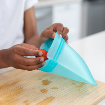 Hands place a strawberry into a Bumkins Silicone Flat Reusable Bag 2 Pack, Blue over a wooden cutting board.
