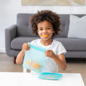 A child with curly hair holds a reusable silicone bag containing sliced oranges. They are smiling with an orange slice in their mouth, sitting at a table in a living room with a gray sofa in the background.
