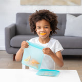 At a table, a child joyfully holds a Bumkins Silicone Flat Reusable Bag 2 Pack, Blue while they smile with an orange slice in their mouth.