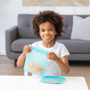 At a table, a child joyfully holds a Bumkins Silicone Flat Reusable Bag 2 Pack, Blue while they smile with an orange slice in their mouth.