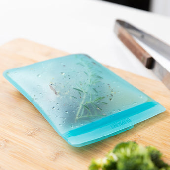 A Bumkins Silicone Flat Reusable Bag 2 Pack, Blue filled with herbs and meat sits on a wood cutting board on a kitchen counter.