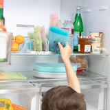 A child reaching into an open fridge to grab a Bumkins Silicone Flat Reusable Bag 2 Pack, Blue among other bags, food, and drinks.