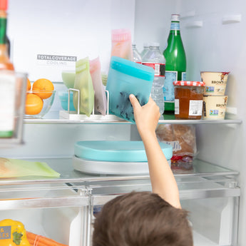 A child reaching into an open fridge to grab a Bumkins Silicone Flat Reusable Bag 2 Pack, Blue among other bags, food, and drinks.