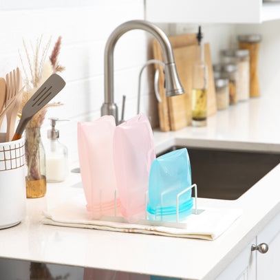 Reusable silicone bags in pink and blue are drying on a rack on a white kitchen counter. A silver faucet is in the background, with jars and kitchen utensils nearby.