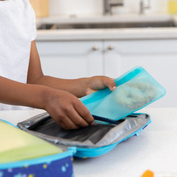 A person packing a Bumkins Silicone Flat Reusable Bag 2 Pack, Blue into an open lunchbox on a kitchen counter.