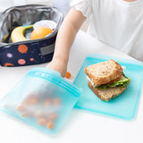 A child's hand reaches for snacks from a Bumkins Silicone Flat Reusable Bag 2 Pack, Blue beside a sandwich, with a lunch bag in the background.