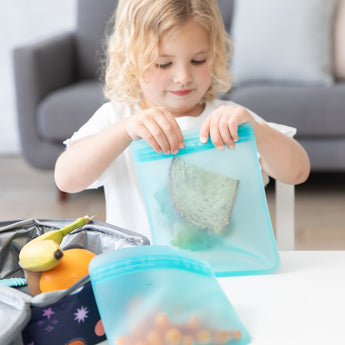 A young child seals a Bumkins Silicone Flat Reusable Bag 2 Pack, Blue with a sandwich at a white table next to a lunchbox with fruit.