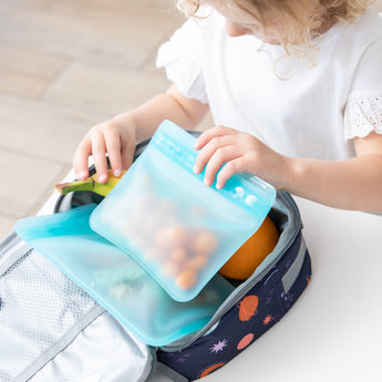 A child packs snacks into an eco-friendly lunchbox at a white table using Bumkins Silicone Flat Reusable Bag 2 Pack, Blue.