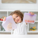 A child happily holds Bumkins Silicone Flat Reusable Bag 2 Pack, Lavender full of snacks, sitting at a white table with shelves behind.