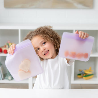A child happily holds Bumkins Silicone Flat Reusable Bag 2 Pack, Lavender full of snacks, sitting at a white table with shelves behind.
