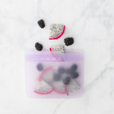 A Bumkins Silicone Flat Reusable Bag 2 Pack, Lavender, filled with fruit slices, lies on a sleek marble surface.