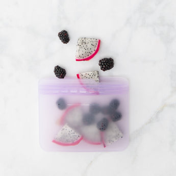 A Bumkins Silicone Flat Reusable Bag 2 Pack, Lavender, filled with fruit slices, lies on a sleek marble surface.