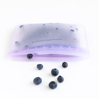 Blueberries spill out of an open Bumkins Silicone Flat Reusable Bag 2 Pack, Lavender on a white background.