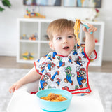 A boy wearing Bumkins Junior Bib: Super Mario™ & Luigi in a high chair looks surprised as he holds spaghetti on a fork from a blue bowl.