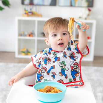A boy wearing Bumkins Junior Bib: Super Mario™ & Luigi in a high chair looks surprised as he holds spaghetti on a fork from a blue bowl.