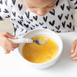 Close-up of a girl in a black & white patterned bib eating soup from a Bumkins Silicone Grip Bowl: Marble with a spoon.