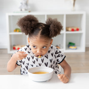 A girl in pigtails wearing a black & white patterned bib eats soup from a Bumkins Silicone Grip Bowl: Marble with a spoon at a white table.
