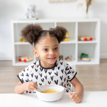 A girl with curly hair wears a black & white bib and eats soup with Bumkins Grip Bowl Essential 4-Piece Mealtime Set: Marble at a table.
