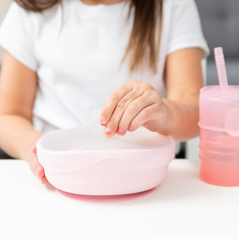 Close-up of a girl lifting a Bumkins Silicone Stretch Lid for Grip Bowl off a pink bowl on a white table next to a pink cup.