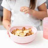 Close-up of a girl lifting a clear lid from a Bumkins Silicone Grip Bowl: Pink with cereal and banana slices on a table next to a pink cup.