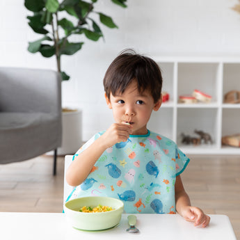 A young boy in a blue bib eats a meal with Bumkins Grip Bowl Essential 4-Piece Mealtime Set: Sage at a white table.
