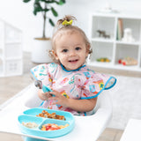 A girl in a high chair smiles, holding Bumkins Spoon + Fork: Blue by a blue divided plate of food with toy-filled shelves behind.
