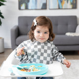 A girl wearing a Bumkins Sleeved Bib: Charcoal Check smirks in a high chair, eating a meal from a blue plate with matching utensils.