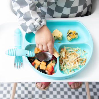 A toddler in a checkered outfit sits in a high chair, using a spoon to eat from a blue divided plate. The plate contains fruit, hash browns, and mini muffins. A matching fork lies beside the plate.