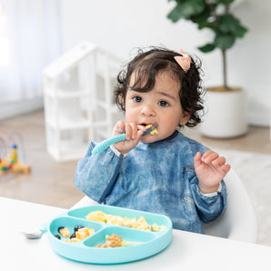 A curly-haired girl enjoys breakfast with Bumkins On-The-Go Mealtime 6-Piece Set: Denim + Blue in a bright playroom.