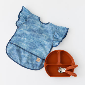 Bumkins Silicone Grip Dish: Clay lays with matching utensils next to a denim-print bib  with ruffle sleeves on a white background.