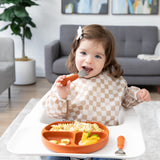 A smiling girl in a tan checkered bib uses Bumkins Spoon + Fork: Clay to eat from a matching plate on a high chair tray.