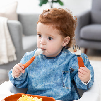 A red-haired girl wearing a blue bib places Bumkins Spoon + Fork: Clay in her mouth as she eats eggs in a cozy living room.