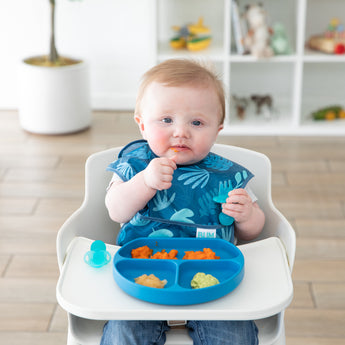 A baby in a blue bib sits in a high chair, practicing self-feeding with Bumkins Silicone Grip Dish: Dark Blue in front of toy-filled shelves.