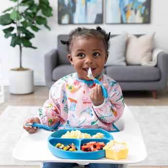 A girl in a pink bib smiles as she eats a meal from Bumkins Silicone Grip Dish: Dark Blue with matching utensils in a cozy living room.